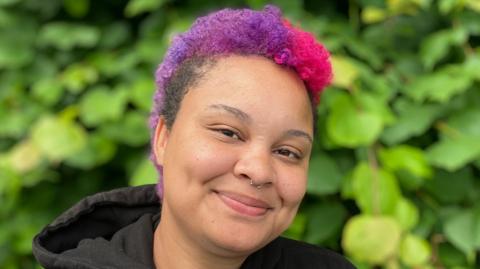 A woman with dyed pink and purple hair smiling in front of a bush