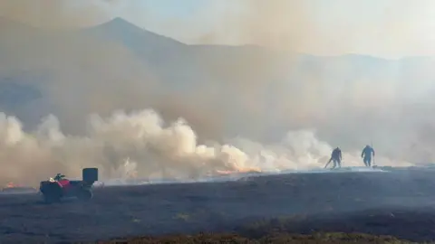 Gamekeepers battle smoke on a field with hills in the distance