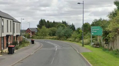 A road curves round to the left with modern houses along the left side. On the right, a grass verge has a sign saying "Welcome to Lightmoor Village".