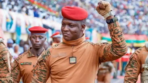 Ibrahim Traoré in beige military fatigues and red beret, surrounded by several soldiers in similar dress, pumps his fist in a stadium of his supporters.