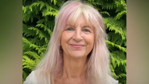 A woman with shoulder length blonde hair with pink highlight smiles as she stands in front of a hedge. She is wearing a white top.