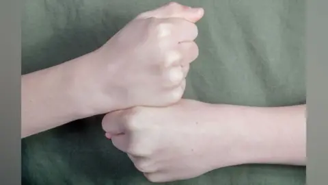 Getty Images A close up of a man's fists against his green-clad torso. His right fist is resting on his left fist in the British Sign Language sign for the letter G.