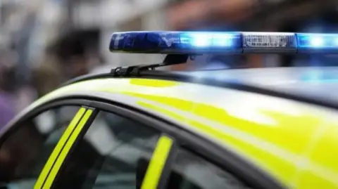 The top of a police car with yellow and white marking and blue and white flashing lights on its roof. The background is blurred.