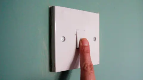 A finger is pressing on a white light switch on a wall. The wall is painted green 