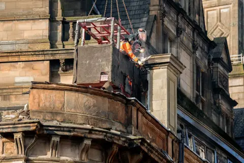 Getty Images Workers in a suspended box lean over the edge and drill at a building facade