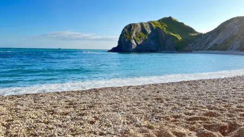 Amy A beautiful blue sea with foamy white waves coming up on to a shingle beach with a headland behind it