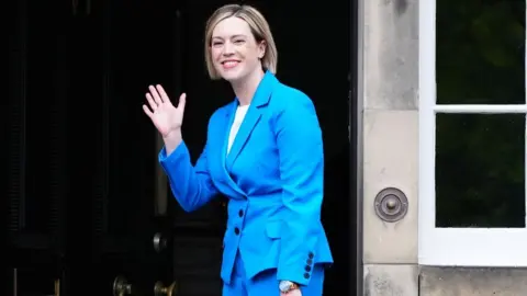 PA Media Jenny Gilruth, who has blonde hair, waves as she stands outside the large black door of a stone building. She is wearing a bright blue suit. 