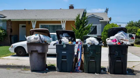 Four overflowing bins stand on the curb in front of a small brown bungalow on a tree-lined street
