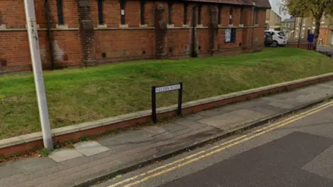 A street view image of a road with double yellow line and a pavement. A street name sign is visible with a grass bank behind it and a red brick building.