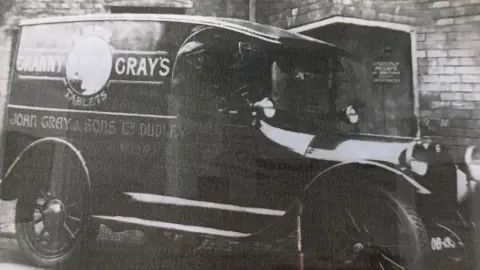 Side view of a black and white old-fashioned car with Granny Grays written on the side as well as John Gray and Songs Limited Dudley. It is parked in front of a brick building. 