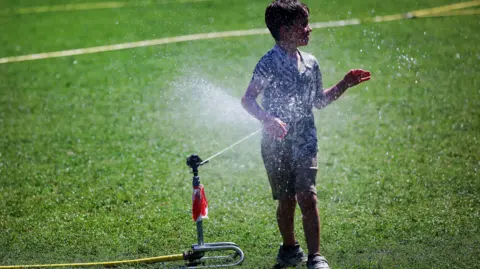 A boy is playing on a green lawn in the water coming from a hosepipe. 