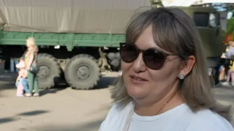 A woman in sunglasses stands in front of an army vehicle