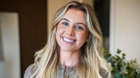 A young woman with blue eyes and long blonde hair looks into camera smiling, her head slightly tilted to the right. She is wearing a grey cardigan with big grey buttons and a gold necklace and earrings