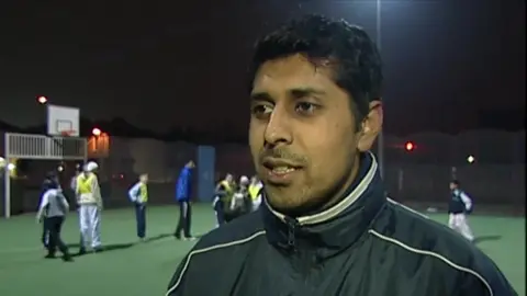 Afrasiab Anwar speaks to camera, he has short dark hair and a small moustache and beard and is wearing a football coaching jacket. In the background young people can be seen playing football on an astro turf pitch.