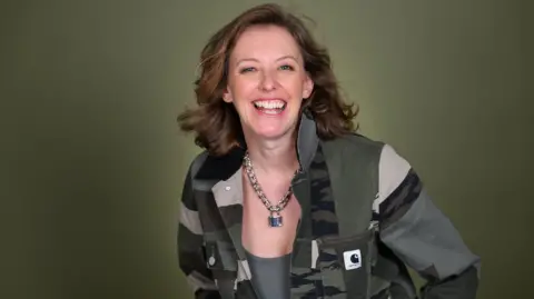 Rosie Wilby A woman with brown hair smiling wearing a khaki jacket in front of a green background. 