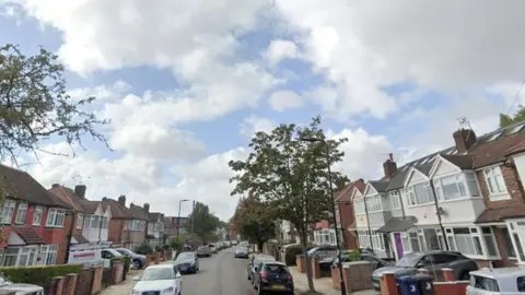 Google Jubilee Road in Perivale, London, with terraced houses on either side of the street and a tree on the right hand side. Cars also line the streets and the sky is blue with white clouds.