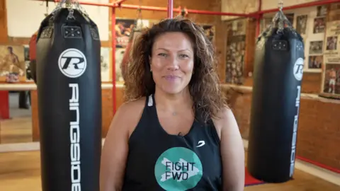 Yurdal Mohammad stands in front of two black punching bags inside a boxing gym, wearing a black Fight Forward vest and smiling at the camera.