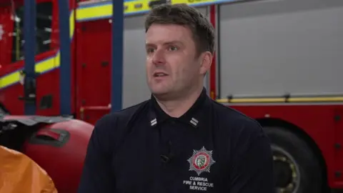 Stuart Forrester, who is clean shaven with short neat fair hair and wearing a navy polo shirt with the Cumbria Fire and Rescue badge on the chest, sits in front of a large red fire engine.