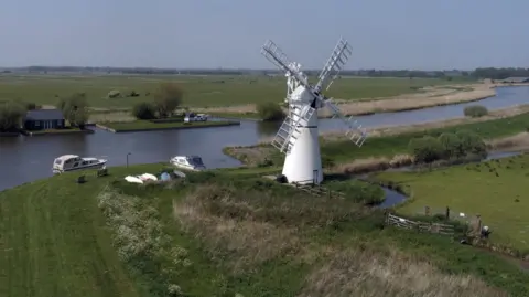 Martin Barber/BBC A white windmill on the banks of the River Thurne, with two day boats on the water, surrounded by Broads flatlands and fields.
