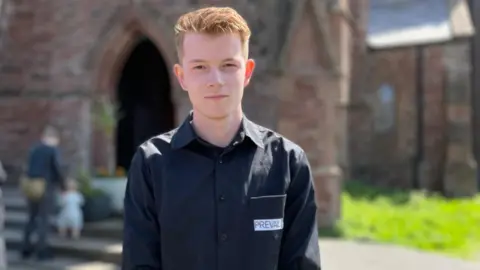 Fabian Kaluza, who has short light brown hair and is wearing a black shirt with Prevail written on the breast pocket. He is standing in front of the cathedral.