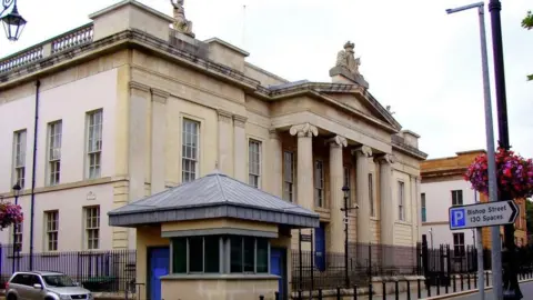 The front of the courthouse in Derry with a silver-coloured car at the left hand side, railings and a security hut, with a parking sign to the right pointing to Bishop Street car park with 130 spaces
