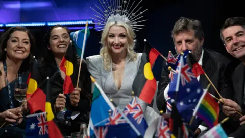 Getty Images Australia's 2019 act Kate Miller-Heidkes surrounded by her team waits for vote results while waiving Australian and Aboriginal flags