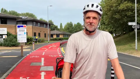 Shariqua Ahmed/BBC Jim wearing a cream T-shirt and a white helmet on his bicycle - along the red cycle path on Thorpe Wood 