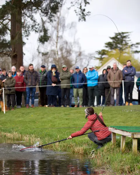 Tweed Salmon Festival A woman in a puffy red jacket and black trousers puts a net out into the water to catch a fish on the end of the rod in her other hand. A small crowd of onlookers is watching from behind a roped off line.