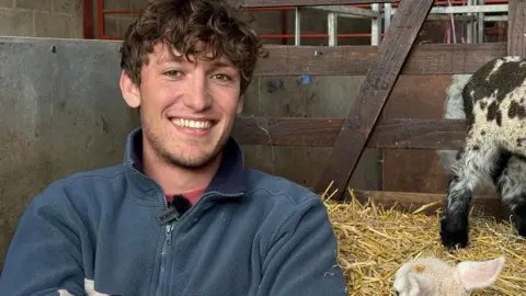 A man with brown curly hair, wearing a blue fleece and smiling as he sits in a lamb pen on some hay.