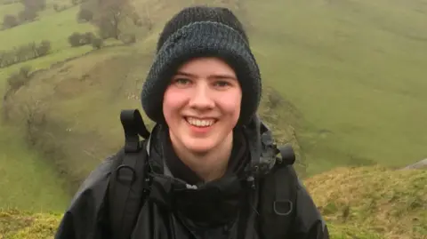 Alex is wearing a black woollen beanie hat, black waterproofs and rucksack, smiling to camera in the rain. Alex is climbing a hill and you can see the green fields below in the background. 