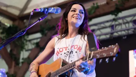 Amy Macdonald performing onstage, wearing a white T-shirt and playing an acoustic guitar.
