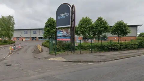 Google A large sign reading 'Total Fitness' - the site where the proposed music academy will be built. The sign sits in front of a large warehouse on an industrial site.
