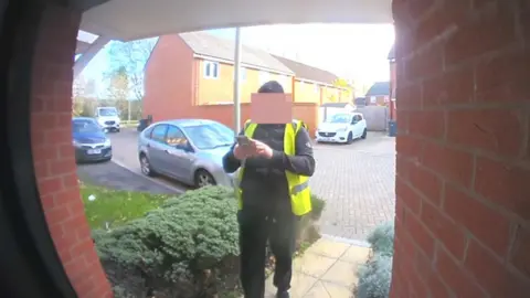 Jean Bryan A picture taken from a doorbell camera showing a man wearing a high-viz jacket and a black hat. He is standing in front of a house taking a picture on his mobile phone.
