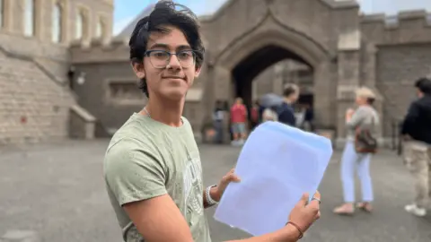 A student holding his GCSE results, which are blurred, standing outside of Vic college.