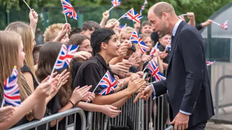 Prince William, dressed in a navy blue suit, is walking past a long crowd of students who are stood behind a steel fence. Many of them wave miniature Union Flags. He has stopped to speak to a male student, who has leaned over the barrier and is clapping his hands.