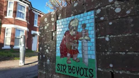 A picture of a scene in Bevois Valley - a mosaic-type picture of a man resembling a knight with a sword can be seen painted on a brick wall. A house can be seen in the background.