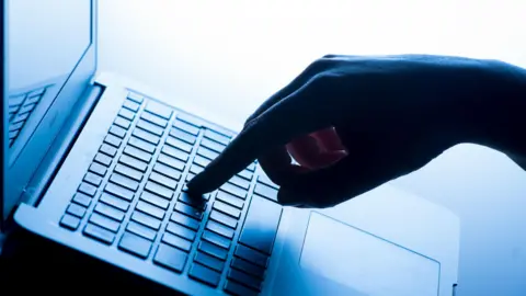 A silhouette of a hand typing on a laptop keyboard. There is a finger on one of the keys and the lighting is low and has a blue tone. The hand is on the right and the laptop is on the left.