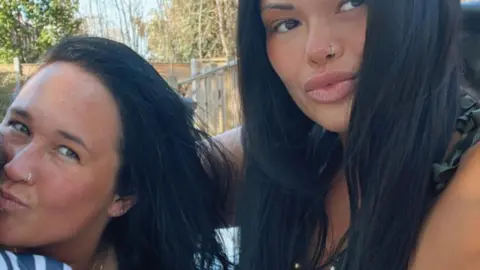 Image of two women with long black hair and nose rings smiling at the camera. In the background can be seen a garden fence, trees and plants.