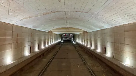 Wienerberger Interior view of a long industrial tunnel with a high, arched brick ceiling and parallel rails running down the centre. Small lights line both sides of the tunnel, illuminating pale stone walls marked with large painted numbers. At the far end, a piece of industrial equipment is visible on the tracks. The image is symmetrical, drawing the eye toward the distant vanishing point and highlighting the scale and length of the structure.