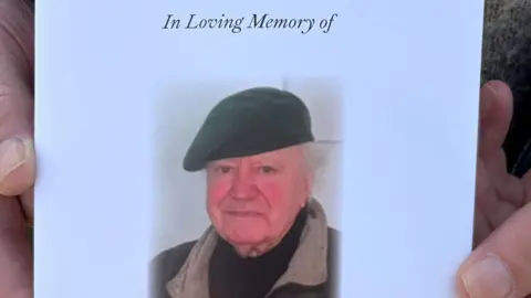 The cover of a funeral programme featuring Tony Martin's photo, with him wearing a beret, black roll neck jumper and jacket with a beige collar. The title says 'In Loving Memory'. The programme is being held by someone's hands in shot.