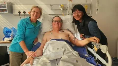 Roland and Shirley Cherry Roland Cherry is lying in a hospital bed with his wife and another woman by his side. He has tube and stickers on his bare chest with the two women either of him. His wife is wearing a blue top and brown trousers. The second woman is in dark top and trousers. All are smiling in the photo.
