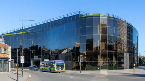 Dark glass curved building reflects the architecture and buildings opposite. A bus has paused at traffic lights. 