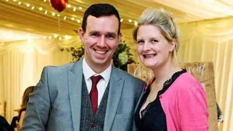 Brain Tumour Research Charlie and Siobhan in an older photo, possibly taken at a function or a wedding. Charlie has short dark hair and wears a grey suit, waistcoat and red tie. Siobhan wears a black blouse and pink cardigan.