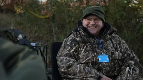 Jamie Niblock Brian Haycock sitting down and smiling at the camera. He is wearing a camouflage style jacket and a green hat.