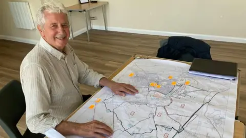 A man is smiling at the camera. He is sitting in front of a map with lots of notes and orange stickers.
