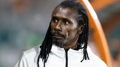 A head shot of Aliou Cisse wearing a white tracksuit top with his dreadlocks hanging loose