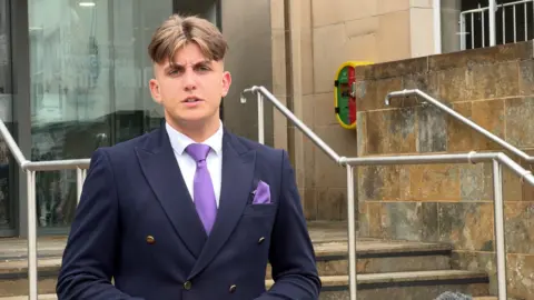 Reform UK leader of Warwickshire County Council, George Finch wears a dark blue double-breasted jacket, white shirt and purple tie and handkerchief in his top pocket. He is standing on steps outside the council house. 