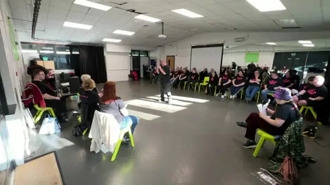 Inside a rehearsal room, a number of performers sit down on green chairs in front of some mirrors. They are all wearing black T-shirts that says "All Stars Theatre Group". One man is standing the middle of the room, holding a microphone and singing. Several people are also sat at a desk.
