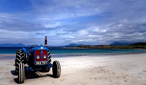 Alan MacRae A blue vintage tractor stands on a wide white-sand beach beside turquoise water. Low rocky islands and dark mountains are visible in the distance beneath a dramatic sky filled with thick grey and blue clouds.