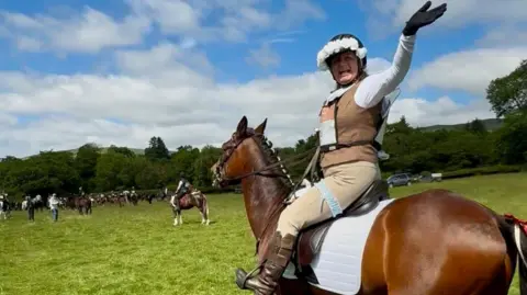 Family photo Safron sat on a brown horse wearing her racing unifrom with a white vale underneath her helmet. She has one hand in the air and smiles at the camera. In the background there are people on horses ready to begin the race. 
